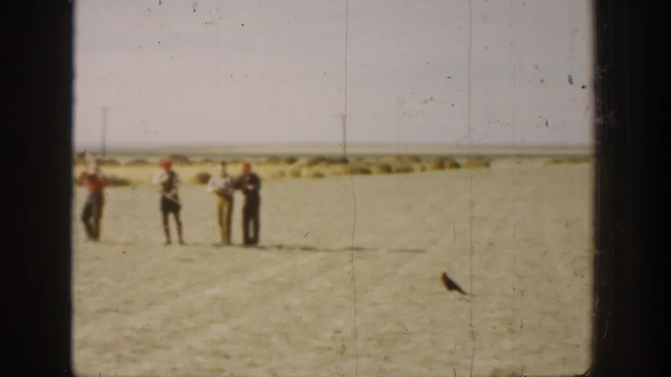 1936 California 8mm frame showing four distant men in a flat field and a dark bird near the lower right edge.
