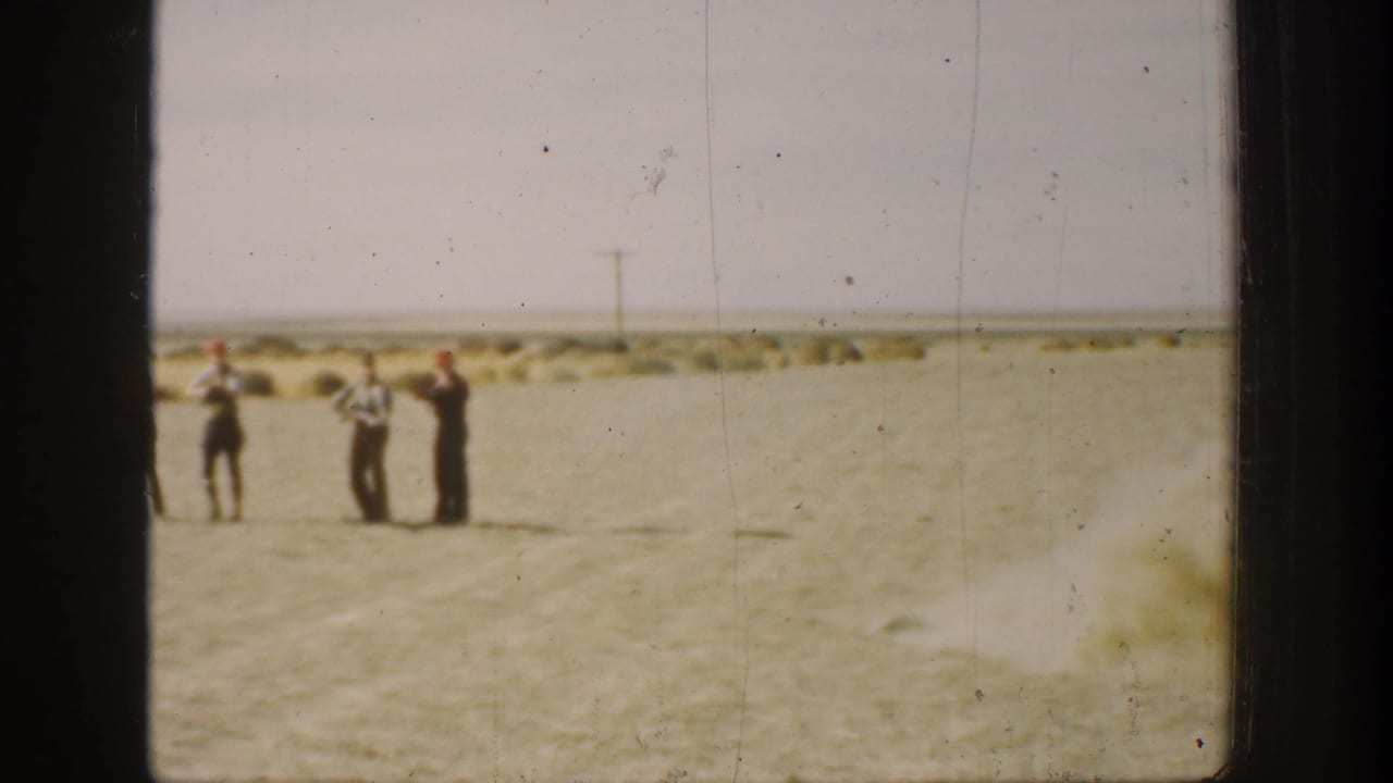 Three men stand apart in the sandy field, with rifles visible near their shoulders.