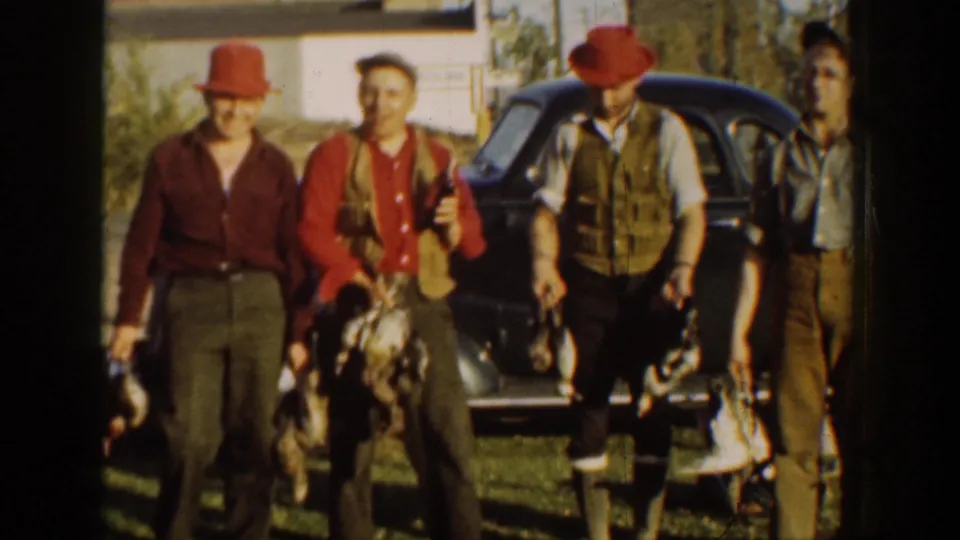 Three adult men in period outdoor clothing pose with game birds beside a dark 1930s automobile in California home-movie footage.