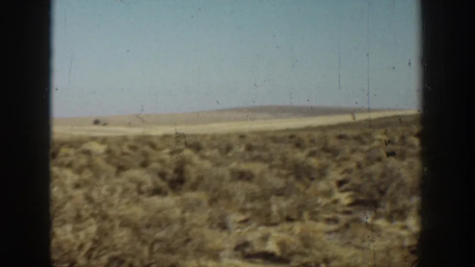 1936 California 8mm frame showing a dry open field with scattered shrubs, low hills, pale blue sky, and worn film edges.
