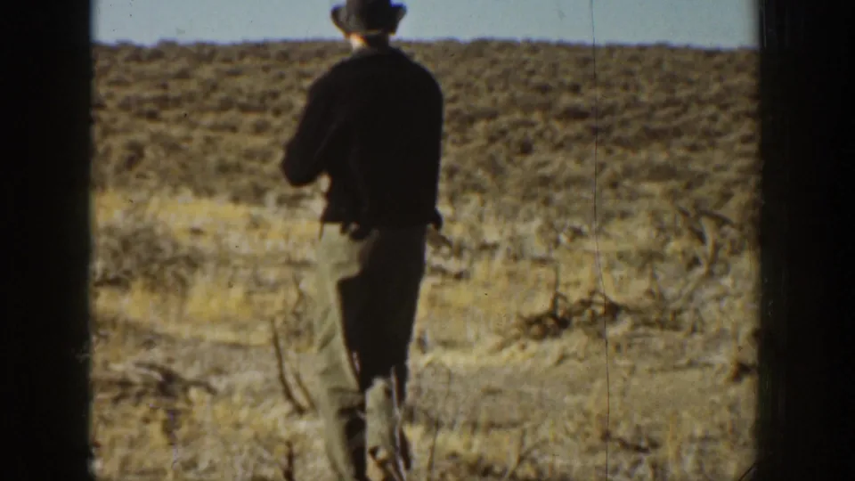 1936 California home-movie frame of a man seen from behind, walking through arid scrubland below a pale sky.