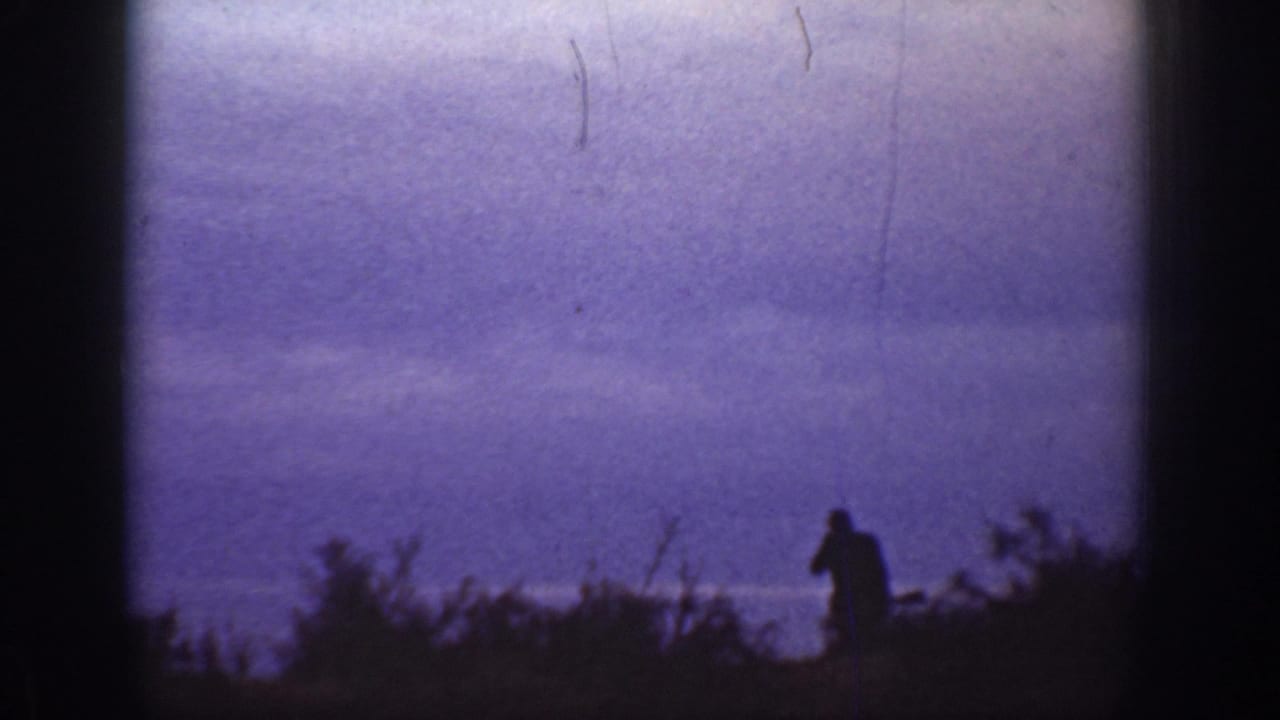 A distant silhouetted person crosses a brushy California field line in low dusk light, with film scratches visible.