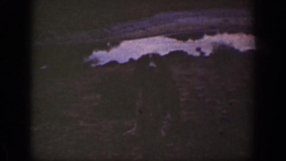 1930s California 8mm frame with a pale ditch or wash edge across dark semi-desert ground and tiny distant figures.