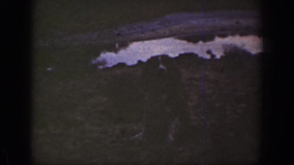 1930s California 8mm frame showing a pale wash or ditch edge, dark scrubby ground, and small indistinct figures below it.