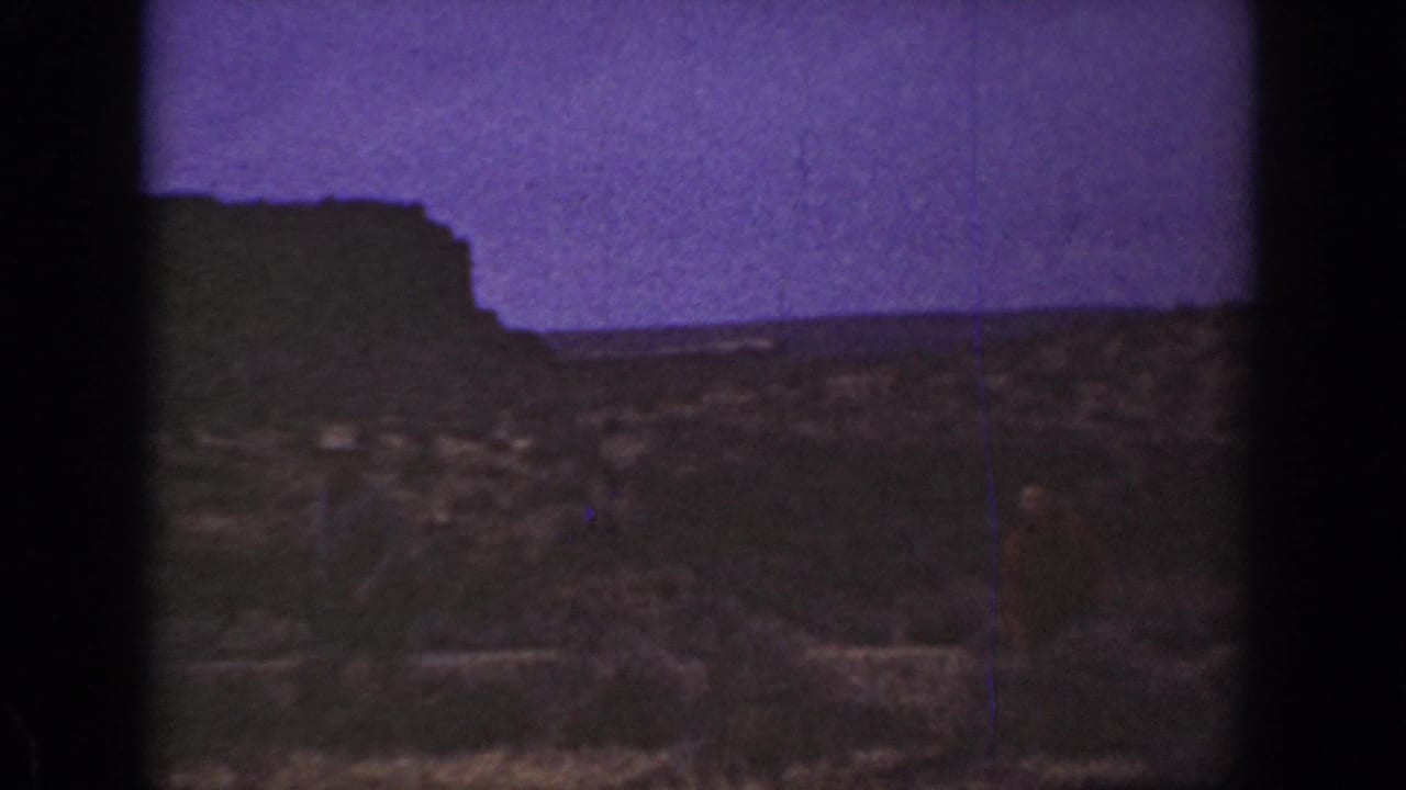 1936 California 8mm frame showing rough arid ground, low vegetation, a dark ridge, and a purple-tinted sky with no clear people visible.