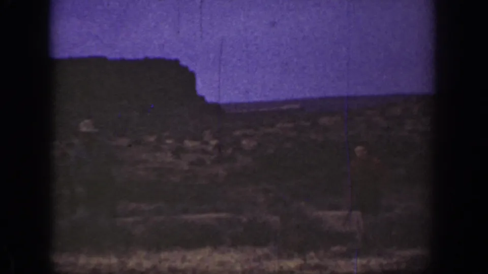 1936 California 8mm frame with rocky desert terrain, sparse vegetation, a dark ridge, and a faint adult figure in the distance.