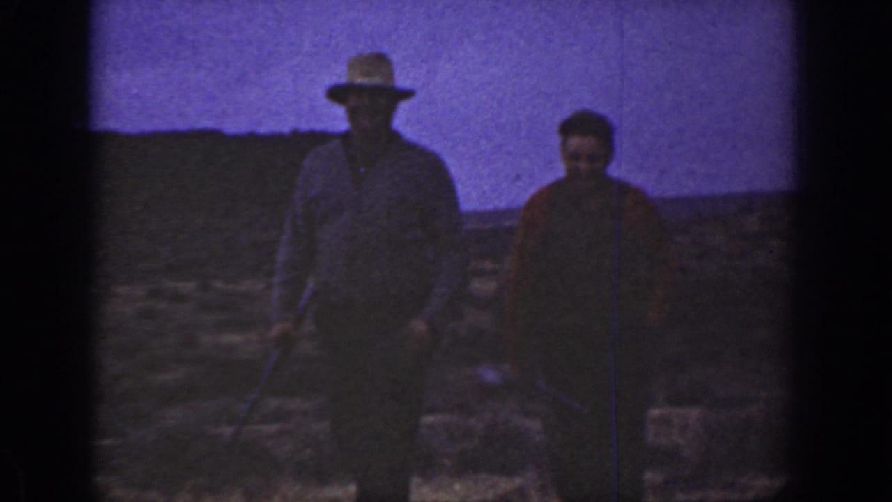 A hat-wearing man and a companion walk toward the camera across rocky open terrain.