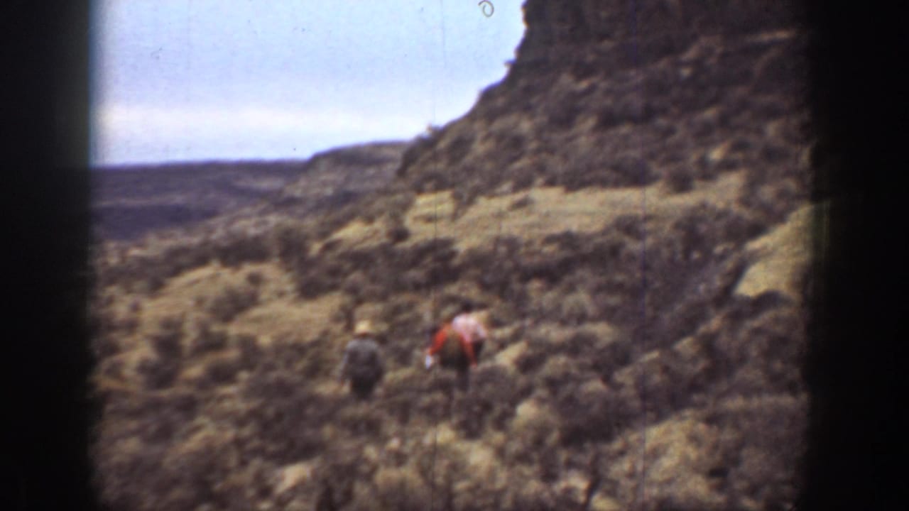 1936 California 8mm frame showing three distant hikers crossing scrub-covered canyon ground beneath a rocky slope.