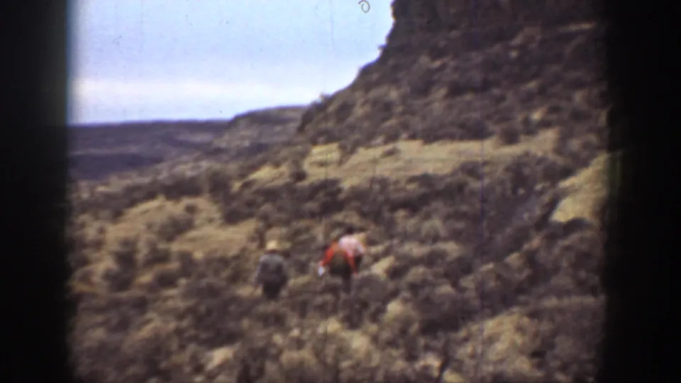 1936 California 8mm frame showing three distant hikers crossing scrub-covered canyon ground beneath a rocky slope.