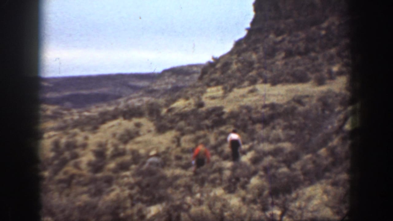 Two hikers continue across scrub near a steep canyon wall.
