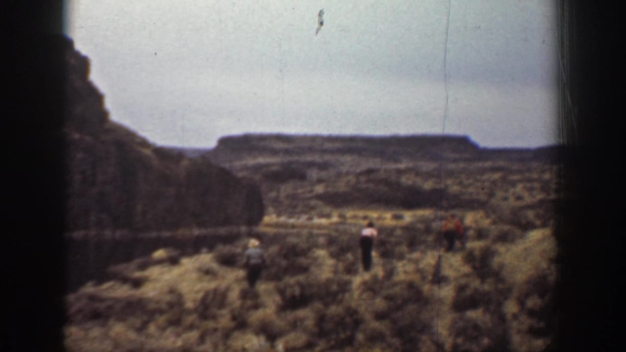 1936 California 8mm frame showing three distant people moving across arid brushland below rocky plateau walls.