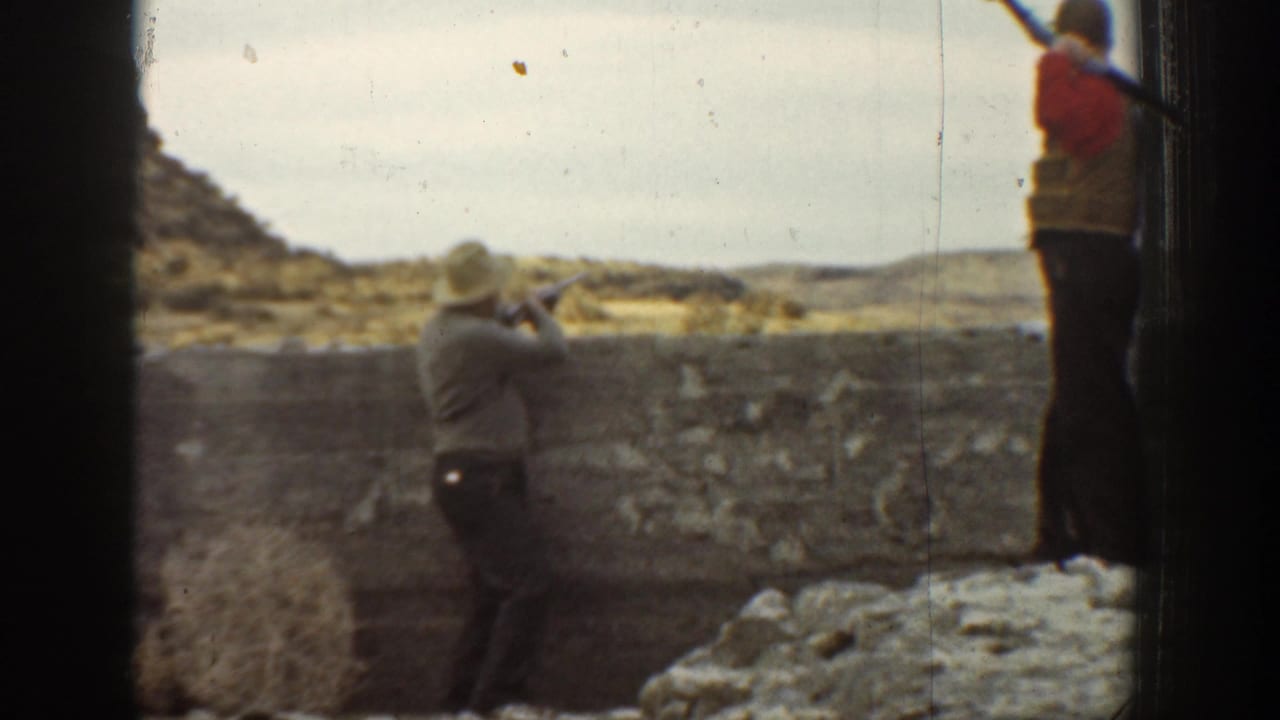 1936 California home-movie frame showing a boy aiming a rifle from behind a stone wall with another boy standing in a dry landscape.