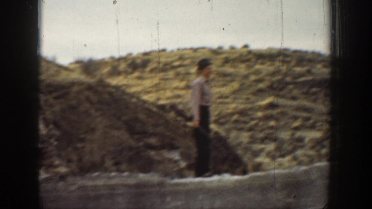 A boy stands near a low stone wall in a dry California landscape, with another figure partly obscured by the film frame edge.