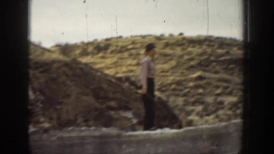A boy stands near a low stone wall in a dry California landscape, with another figure partly obscured by the film frame edge.