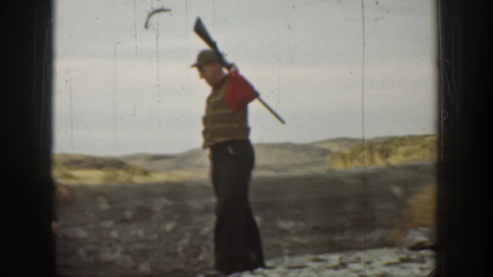 1936 California frame of a boy in a cap and red vest holding a rifle or long object over his shoulder beside a stone wall.