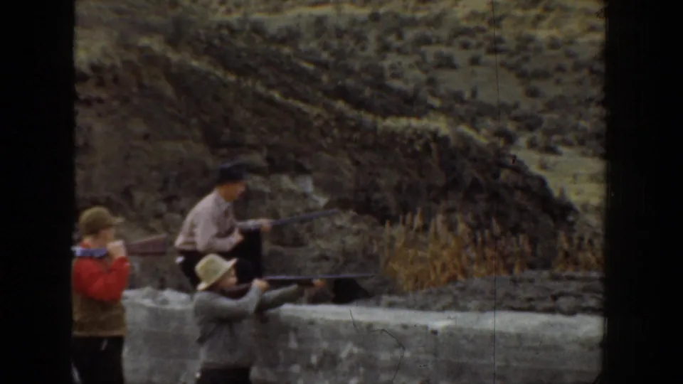 Boys gather beside a low wall in 1936 California, with rifles visible and dry hills rising behind them.