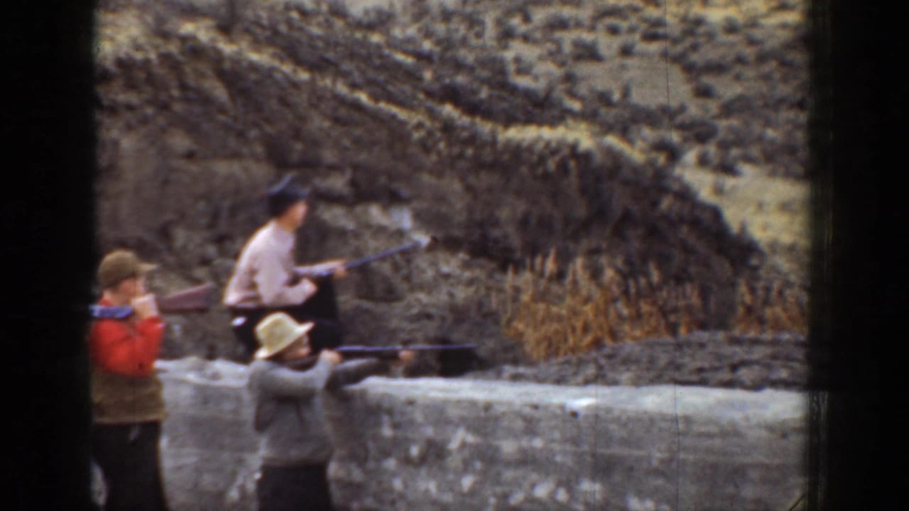 1936 California 8mm frame showing three men in hats aiming rifles beside a low wall with dry brush and hillside behind them.