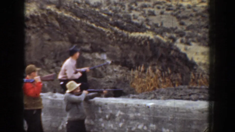 1936 California 8mm frame showing three men in hats aiming rifles beside a low wall with dry brush and hillside behind them.