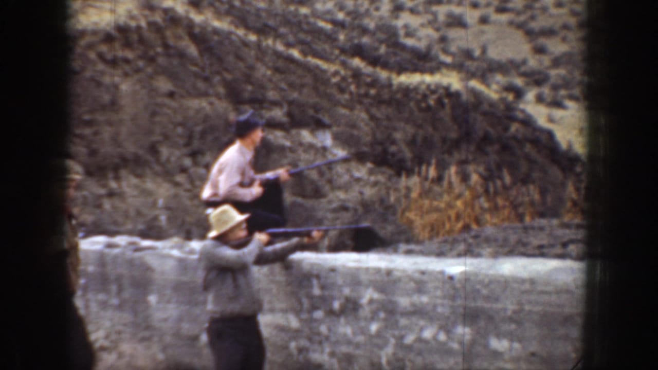 1936 California 8mm frame with two men in hats aiming rifles toward the right beside a low wall and dry hillside.