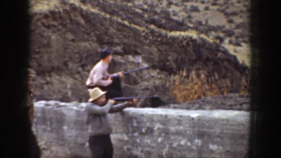 1936 California 8mm frame with two men in hats aiming rifles toward the right beside a low wall and dry hillside.