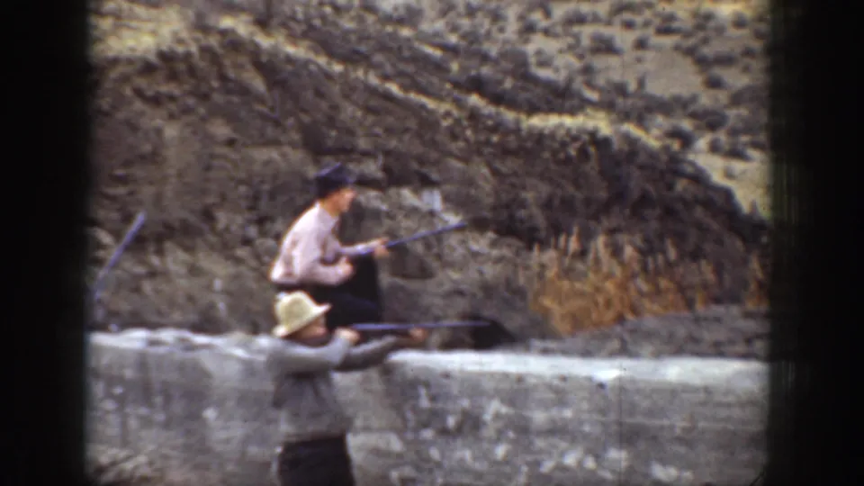 1936 California 8mm frame showing two men with rifles near a low wall, with dry hillside terrain behind them.