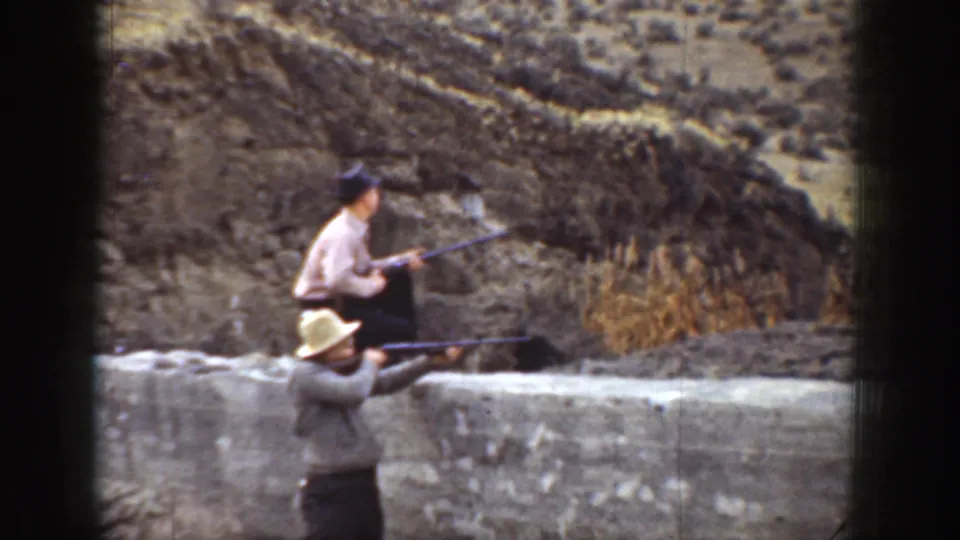 1936 California 8mm frame of two men in hats holding rifles beside a low wall in a dry hillside setting.
