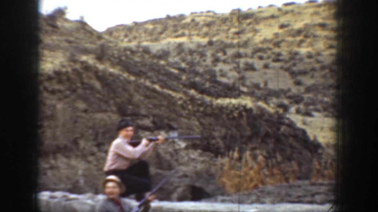 In 1936 California home-movie footage, a man crouches with a rifle behind a low stone wall in an arid hillside setting.
