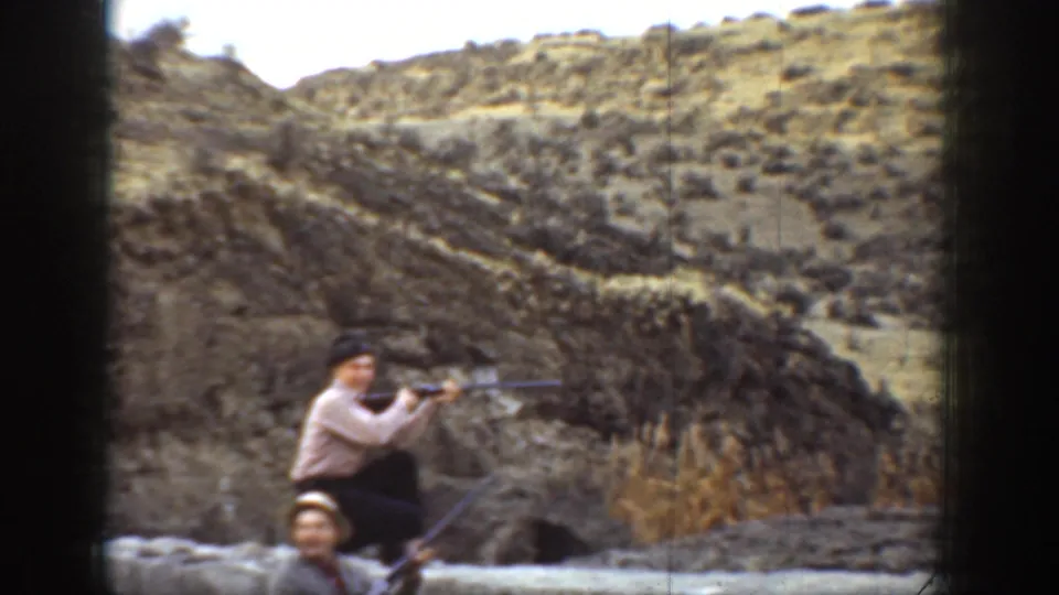 In 1936 California home-movie footage, a man crouches with a rifle behind a low stone wall in an arid hillside setting.