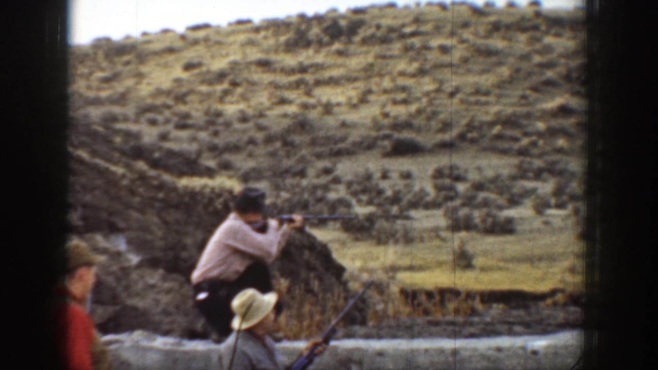 Two men with rifles appear near a low wall below dry hills.