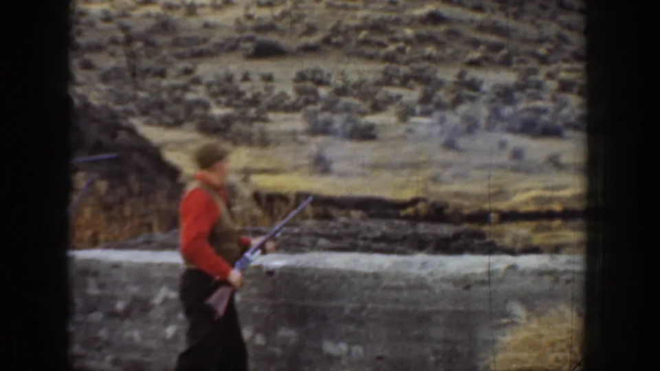 A man in a red shirt and vest carries a rifle beside a low wall, with dry California hills behind him.