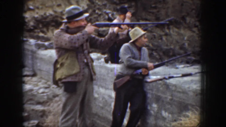 1936 California home-movie frame showing men in period hats aiming long rifles behind a concrete wall at an outdoor range.