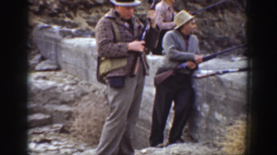 1936 California home-movie frame with men holding rifles beside a low concrete wall in a dry, rocky outdoor range area.