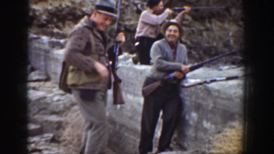 1936 California home-movie frame of men holding long rifles and smiling near a concrete wall in an arid outdoor range setting.