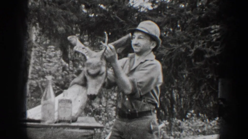 In this 1939 Chelan outdoor home-movie frame, a man in a hat handles a mounted deer head beside tabletop bottles and trees.