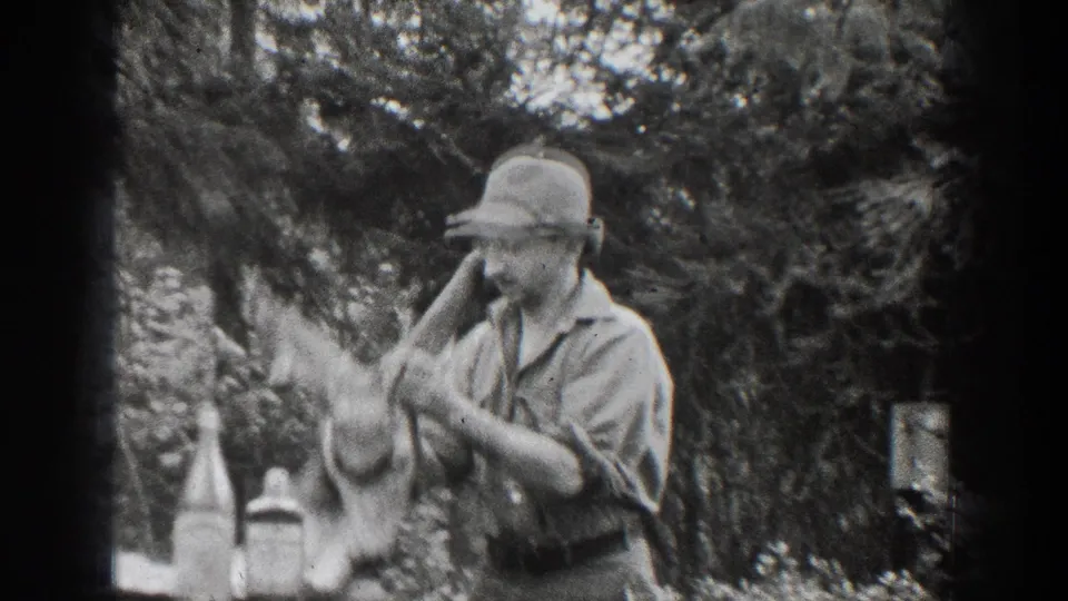 1939 Chelan home-movie frame showing the man leaning toward the mounted deer head, with trees and tabletop objects behind the soft film grain.