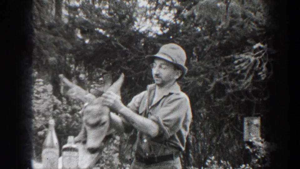 In 1939 Chelan, Washington footage, a man in a hat and work shirt handles a deer head with antlers in a wooded setting.