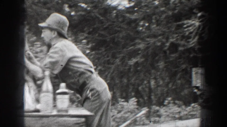 The man appears in profile, leaning toward the deer head on or near a table in the wooded Chelan home-movie scene.