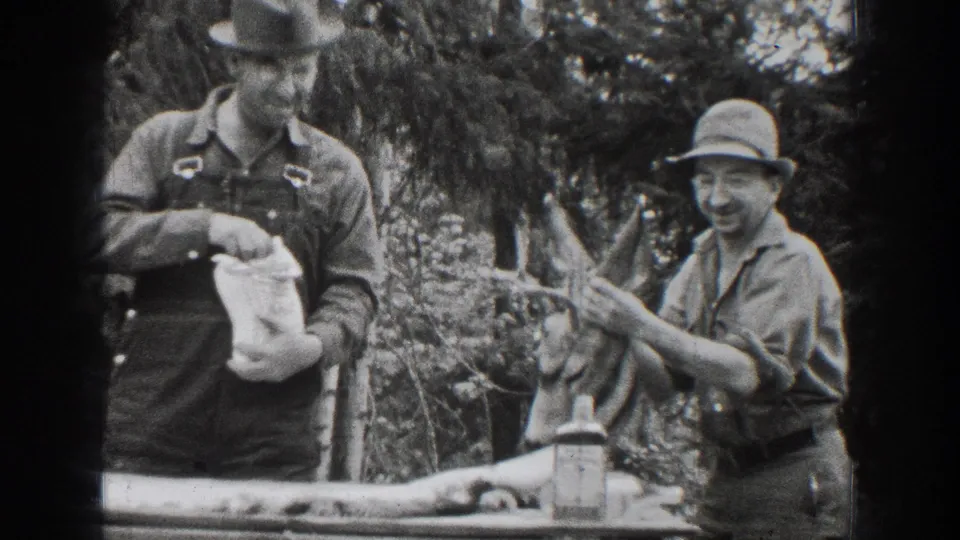 Two adult men appear in the wooded 1939 Chelan footage, with one laughing as the other stands near the table and deer head.