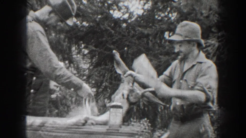 A man in a hat holds the antlered deer head as another adult man is partly visible in the Chelan, Washington home-movie frame.