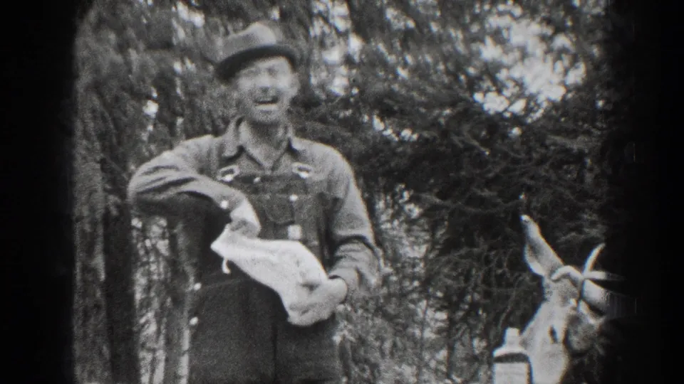 A smiling man in overalls faces the camera in wooded 1939 Chelan footage, with a deer at the right edge.