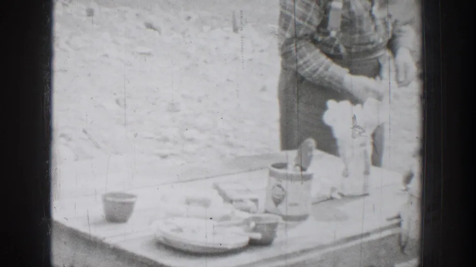 1939 Chelan, Washington home-movie frame showing a camp table with dishes, tins, and a man partly visible beside outdoor meal supplies.