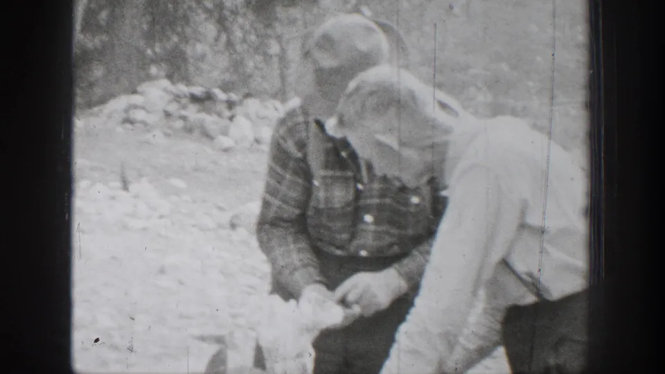 1939 Chelan, Washington frame showing two hunters in brimmed hats and work clothes handling food during a forest meal preparation.