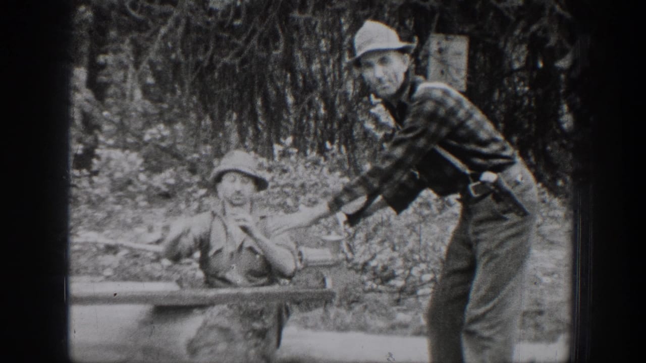 Chelan, Washington 1939 home-movie frame showing two adult men beside a log work surface, with a metal basin and wooded brush behind them.