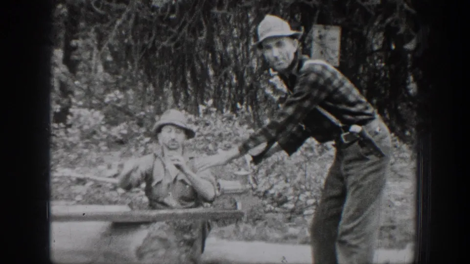 Chelan, Washington 1939 home-movie frame showing two adult men beside a log work surface, with a metal basin and wooded brush behind them.