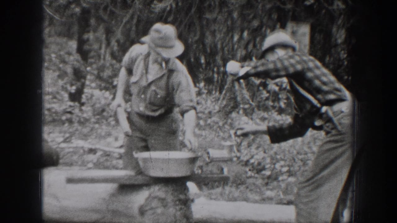 One man bends over a metal basin while another reaches toward the log bench.