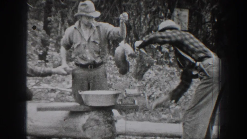 Chelan, Washington 1939 home-movie frame showing a man lifting a metal container while his companion bends near the log work area.