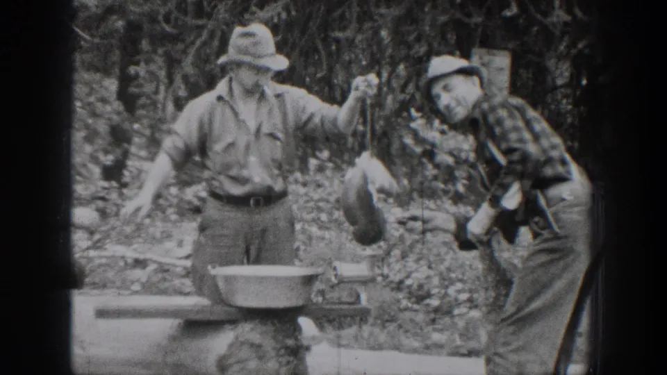 Chelan, Washington 1939 frame of two men near a log bench, one holding a metal container and the other leaning toward the camera.