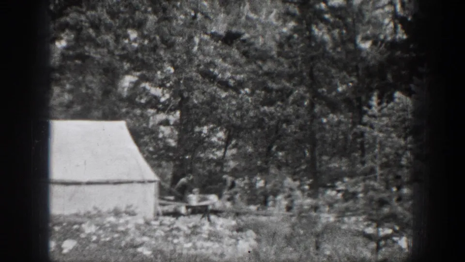 1939 Chelan, Washington camp scene with a white canvas tent and a man working near a small table in dense woods.