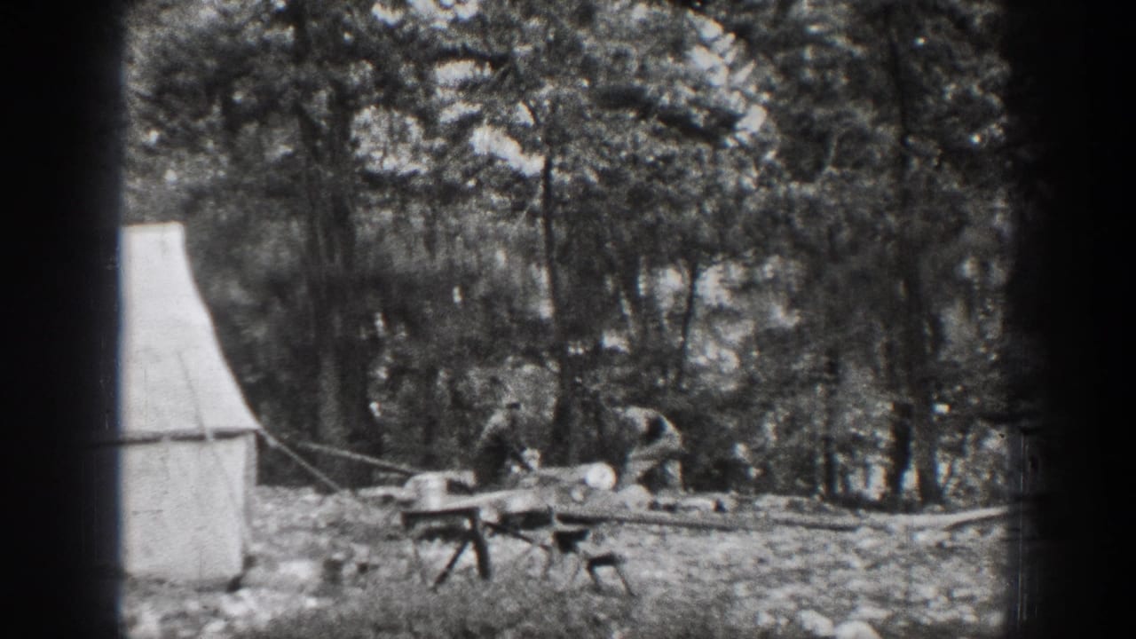 1939 Chelan home-movie frame showing men near a canvas tent, rough tables, and cut lumber in a forest clearing.
