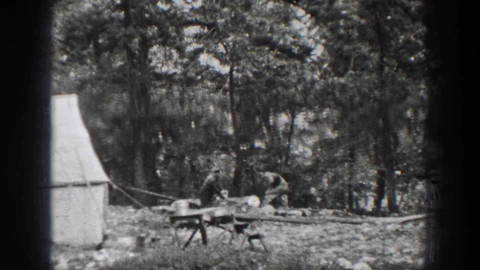 1939 Chelan home-movie frame showing men near a canvas tent, rough tables, and cut lumber in a forest clearing.